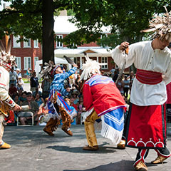 Colour photograph of four dancers wearing their traditional clothing of leather or fabric. They are all also wearing moccasins and a feather headdress on their heads.