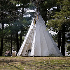 Photographie couleur d'un wigwam près d'arbres. Trois personnes sont assises juste à côté.