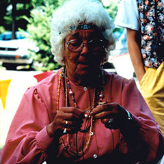 Photographie couleur d'une dame aux cheveux blancs, courts et frisés, portant une chemise rose et plusieurs colliers de perles colorées.
