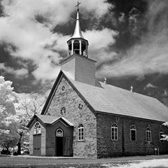 Black and white photograph of a church made of stones and its bell tower. Large white clouds are also visible.