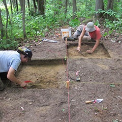 Colour photograph of two trainee archaeologists digging a site in a wooded area.