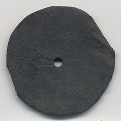 Photographie couleur d'un disque d'ardoise grise d'un diamètre de 10 cm, percé au centre. Le disque est gravé de symboles en forme de chevron et de lignes doubles.