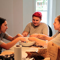 Colour photograph of three young trainee archaeologists cleaning artifacts with various tools on top of a bowl of water.