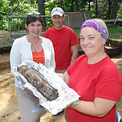 Colour photograph of two women and a man. The first woman holds a charred post in her hands. The two other persons are smiling behind her.