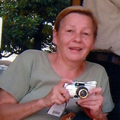 Photographie couleur d'un femme souriante, portant un t-shirt vert. Elle tient dans ses mains un appareil photo.